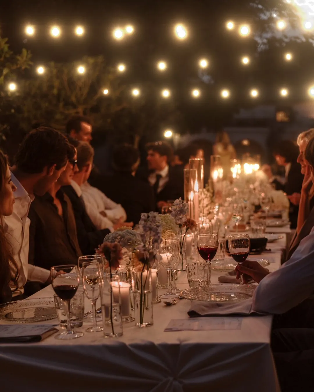 Mesa larga de boda al aire libre, con velas, flores y guirnaldas de luces encendidas